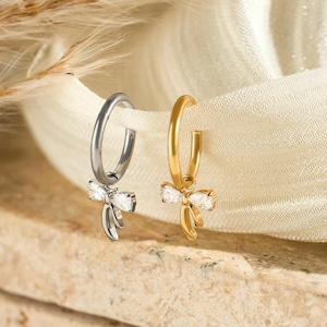 May include: Two hoop earrings, one silver and one gold, each with a bow-shaped charm adorned with clear gemstones. The earrings are displayed on a neutral-toned surface with soft, flowing fabric in the background.