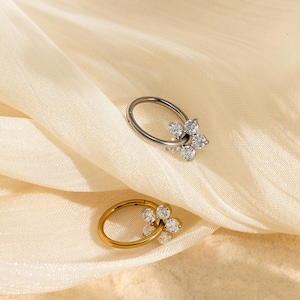 May include: Two nose rings with flower designs. One ring is silver-colored, and the other is gold-colored. Each ring features a small flower made of clear, sparkling stones. The rings are displayed on a cream-colored, draped fabric.
