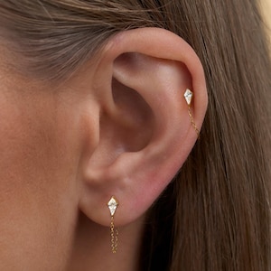 May include: A close-up of an ear with two gold earrings. Each earring has a diamond-shaped design with a small chain connecting them. The earrings are set with small, clear gemstones, creating a delicate and elegant look.