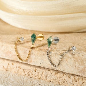 May include: Two pairs of earrings in gold and silver. Each earring has a green gemstone and a small chain leading to a small, clear stone. The earrings are displayed on a textured, light-coloured surface.