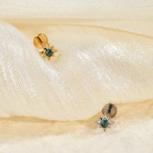 May include: Two star-shaped stud earrings. One earring is gold-toned, the other is silver-toned. Each earring features a small, dark green gemstone at its centre. The earrings are displayed on a cream-coloured, textured fabric.