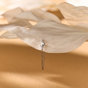 May include: A silver-toned earring featuring a kite-shaped clear gemstone set in a metal frame with a delicate dangling chain attached to the base. The piece is positioned against a large, smooth, off-white petal with a soft, warm-toned background.