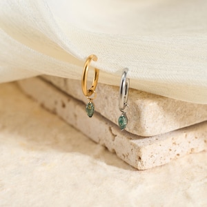 May include: A pair of hoop earrings, one gold-coloured and one silver-coloured, each featuring a small, green, teardrop-shaped charm. The earrings are presented on a textured, light-coloured surface with a soft, white fabric. Jewellery.