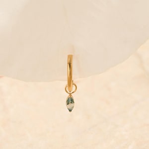 May include: A gold-colored hoop earring with a small, teardrop-shaped charm. The charm is a mix of green and white, adding a touch of color to the minimalist design. The earring is set against a soft, off-white background.