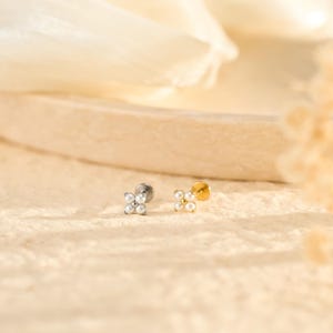 May include: Two small, flower-shaped earrings with a stud backing. One earring is silver-toned, the other is gold-toned. Each earring features four small, round, white pearls arranged in a floral pattern. The earrings are displayed on a textured, light-colored surface.