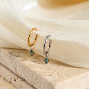 May include: Two hoop earrings, one gold-toned and one silver-toned, each with a small, oval, green gemstone pendant. The earrings are displayed on a textured, light-colored surface with a soft, white fabric in the background.