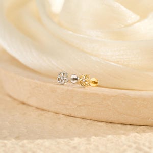 May include: Two flower-shaped stud earrings. One is silver-coloured, the other is gold-coloured. Each earring features a small, clear gemstone in the centre of the flower design. The earrings are displayed on a light brown surface.