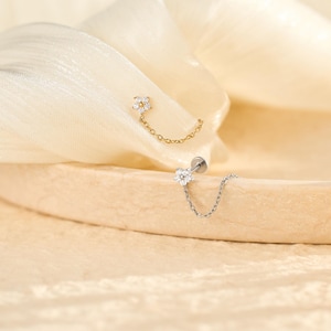 May include: Two flower-shaped stud earrings with attached delicate chains. One earring is gold-toned with clear stones, and the other is silver-toned with clear stones. Each features a flat back post and a small floral charm connected by a chain.