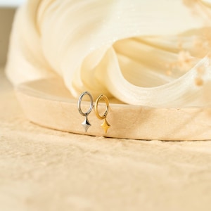 May include: Two hoop earrings, one silver and one gold, each with a dangling star charm. The earrings are displayed on a light wooden surface against a soft, neutral background. The star charms add a touch of elegance.