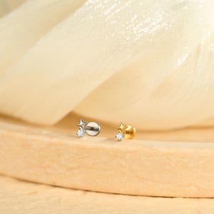 Könnte beinhalten: Zwei kleine sternförmige Ohrringe. Einer ist silberfarben, der andere goldfarben. Jeder Ohrring hat einen kleinen, klaren Edelstein. Die Ohrringe werden auf einer hellbraunen Oberfläche mit einem weichen, cremefarbenen Stoff im Hintergrund präsentiert.