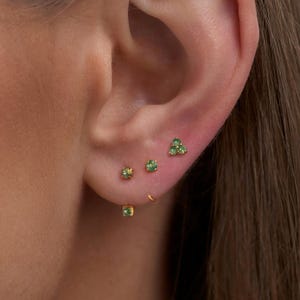 May include: A set of gold stud earrings featuring small green gemstones. The collection includes three individual round green stone studs and one cluster stud composed of three small green stones arranged in a triangular formation.