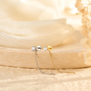 Könnte beinhalten: Zwei Ohrstecker mit einer hängenden Kette. Ein Ohrring ist silberfarben mit einem klaren, runden Edelstein, der andere goldfarben mit einem ähnlichen Edelstein. Die Ohrringe werden auf einer hellbraunen, strukturierten Oberfläche präsentiert.
