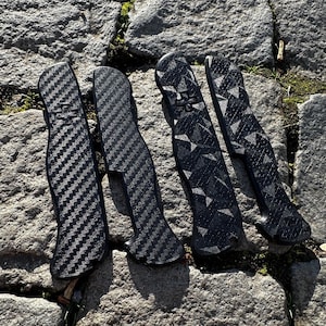 Könnte beinhalten: Vier schwarze Taschenmesser mit verschiedenen gemusterten Griffen. Die Griffe sind aus Carbonfaser.