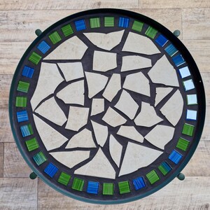 Peut inclure: Une table ronde en métal avec un plateau en mosaïque composé de morceaux de pierre blanc cassé irréguliers au centre. La bordure présente des carreaux de verre carrés verts et bleus alternés sur un joint sombre, dans un cadre en métal vert.