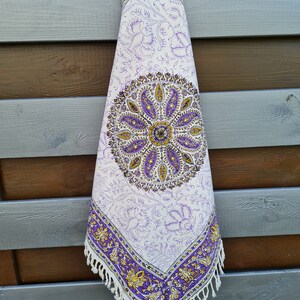 Könnte beinhalten: Eine weiße Baumwolltischdecke mit einem Paisley-Muster in Lila und Gold. Die Tischdecke hat einen Fransenrand und hängt an einer Holzwand.