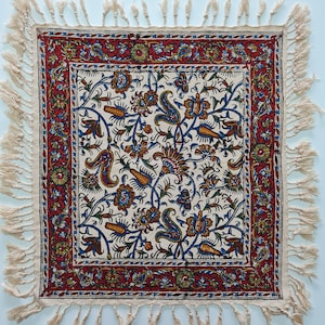 Op de afbeelding: Een witte stoffen tafelkleed met een rood, blauw en groen bloemenpatroon. Het tafelkleed heeft een franje rand.