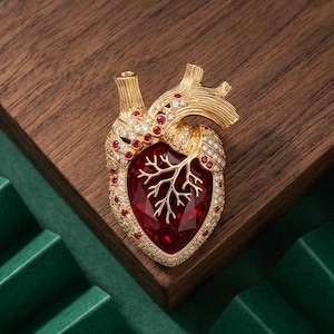 May include: A gold-toned anatomical heart brooch featuring a large, faceted deep red central stone. The surface is embellished with small, clear white crystals and tiny red stones, with a gold branch-like structure overlaying the red center.