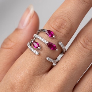 May include: A silver ring with three marquise-cut ruby gemstones and encrusted with small white diamonds. The ring wraps around the finger with an open design, showcasing the gemstones.