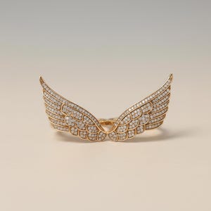 Puede incluir: Un anillo dorado con forma de alas de ángel, incrustado con numerosas piedras preciosas pequeñas y transparentes. Las alas están extendidas, con detalles de plumas. El anillo está sobre un fondo neutro, resaltando el diseño de la joya.