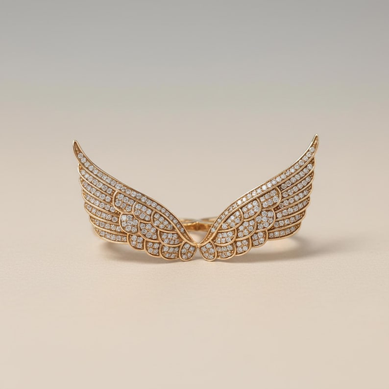 May include: A gold-colored ring shaped like angel wings, encrusted with numerous small, clear gemstones. The wings are spread wide, with detailed feathering. The ring is set against a neutral background, highlighting the jewelry's design.