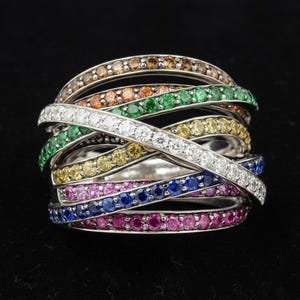 Puede incluir: Un anillo multibanda con un diseño de bandas plateadas. Las bandas están adornadas con una variedad de piedras preciosas en colores como marrón, naranja, verde, blanco, amarillo, rosa, azul y morado. El anillo está sobre un fondo negro.