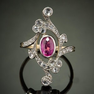 Può includere: Un anello antico con una gemma rosa ovale al centro, circondata da un design decorativo e vorticoso di gemme chiare. L'anello ha una fascia d'oro e un'incastonatura ornata, che riflette la luce.