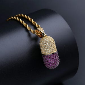 Può includere: Una collana con pendente dorato con un ciondolo a forma di pillola. Il ciondolo è incastonato con piccole pietre scintillanti, con una sezione superiore di pietre chiare e una sezione inferiore di pietre rosa intenso. La catena ha un design a corda ritorta.
