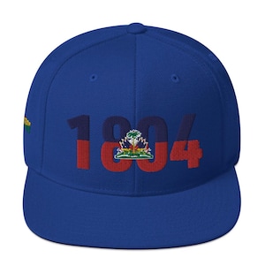 Pode incluir: Boné de beisebol azul royal com o ano "1804" bordado em vermelho, com o "0" apresentando um brasão haitiano. O boné tem uma aba plana e um ajuste estruturado. O lado esquerdo do boné tem um pequeno desenho bordado.