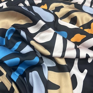 Könnte beinhalten: Seidiger Stoff mit abstraktem Muster. Das Design zeigt schwarze, weiße, blaue, beige und orangefarbene Formen. Der Stoff ist drapiert, wodurch seine Textur und der Fluss des Musters hervorgehoben werden. Geeignet für Kleidung oder Inneneinrichtung.