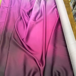 Könnte beinhalten: Ein Stoffballen mit einem lebendigen Farbverlauf, der von tiefem Lila zu hellem Pink übergeht. Der Stoff ist drapiert und zeigt seine glatte Textur und seinen Glanz. Auf der rechten Seite ist eine Rolle weißen Stoffes zu sehen.