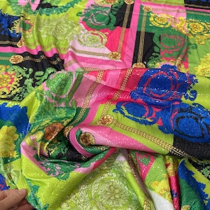 Peut inclure: Un tissu coloré avec un motif géométrique rose, vert, bleu et or. Le tissu est recouvert de paillettes et a une finition brillante.