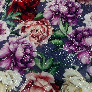 Può includere: Primo piano di un tessuto floreale con grandi fiori colorati. I fiori, in tonalità di rosso, rosa, viola e crema, sono disposti su uno sfondo blu scuro. Il tessuto è impreziosito da paillettes sparse, che aggiungono un effetto scintillante. Il design ricorda un giardino.