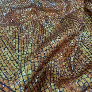 May include: Close-up of a textured fabric with a repeating square pattern. The fabric has a warm colour palette of orange, brown, and gold, with a subtle shimmer. The pattern is outlined in black, creating a grid-like effect.