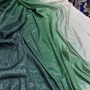 Peut inclure: Un tissu drapé avec un dégradé de verts, passant d'un vert foncé à une teinte pâle, presque blanche. Le matériau a une texture scintillante et à paillettes, captant la lumière et créant un effet étincelant. Le tissu semble doux et fluide.