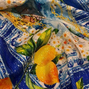 Pode incluir: Close-up de um tecido com um design vibrante com limões, folhas verdes e um padrão azul e branco. O tecido tem uma textura brilhante e com lantejoulas, com detalhes de corrente dourada e detalhes florais. O tema geral é um design de inspiração italiana.