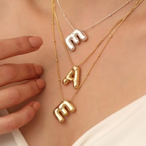 Puede incluir: Tres collares de oro con colgantes de iniciales. El collar superior tiene un colgante de 'M' de plata, el collar del medio tiene un colgante de 'A' de oro y el collar inferior tiene un colgante de 'M' de oro.