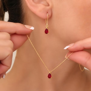 Puede incluir: Un collar de oro y un pendiente de oro a juego con un colgante de piedra roja en forma de lágrima. El collar tiene una cadena de oro fina y el pendiente está diseñado en estilo colgante con un gancho, ambos con una piedra roja vibrante.