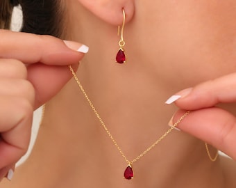 May include: A gold necklace and a matching gold earring featuring a teardrop-shaped red gemstone pendant. The necklace has a delicate gold chain, and the earring is designed as a drop style with a hook, both showcasing a vibrant red stone.