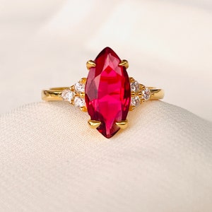 Peut inclure: Une bague dorée ornée d'une grande pierre rouge rubis de forme marquise au centre. La bague est ornée de plus petites pierres précieuses claires de chaque côté de la pierre principale, créant un effet scintillant. La bague est posée sur une surface blanche.