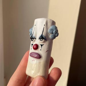 Puede incluir: Figura de cerámica de un payaso triste. El payaso tiene una cara blanca con lágrimas negras, una nariz roja y una boca morada. Tiene pelo azul y está pintado sobre una base rectangular blanca.