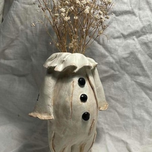 Puede incluir: Un jarrón de cerámica de color blanco roto esculpido a mano con forma de figura, que presenta un cuello con volantes, tres botones circulares negros en la parte delantera y mangas acampanadas. Contiene flores secas de paniculata.