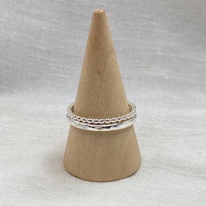 May include: Three silver rings stacked on a wooden ring holder. The top ring is a beaded ring, the middle ring is a hammered ring, and the bottom ring is a plain band.