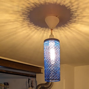 Puede incluir: Una lámpara colgante de malla de alambre azul cuelga de un techo blanco. La lámpara es cilíndrica y tiene una bombilla blanca en su interior.