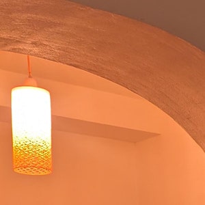 May include: A hanging light fixture with a textured, cylindrical shade. The shade is a light orange color and is illuminated with a warm, white light.