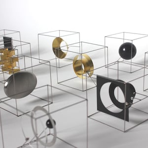 Può includere: Una collezione di cubi in filo metallico contenenti varie forme geometriche, tra cui dischi dorati, una sfera nera, un disco argentato e un quadrato nero con un ritaglio circolare, disposti in una griglia su una superficie piana.