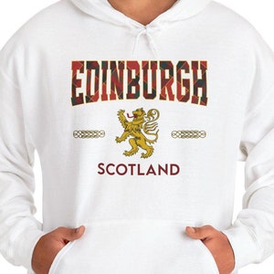 Puede incluir: Sudadera con capucha blanca con la palabra "EDINBURGH" en un diseño de cuadros escoceses en el pecho. Debajo, un emblema de león dorado y la palabra "SCOTLAND" en burdeos. La sudadera tiene un bolsillo delantero.