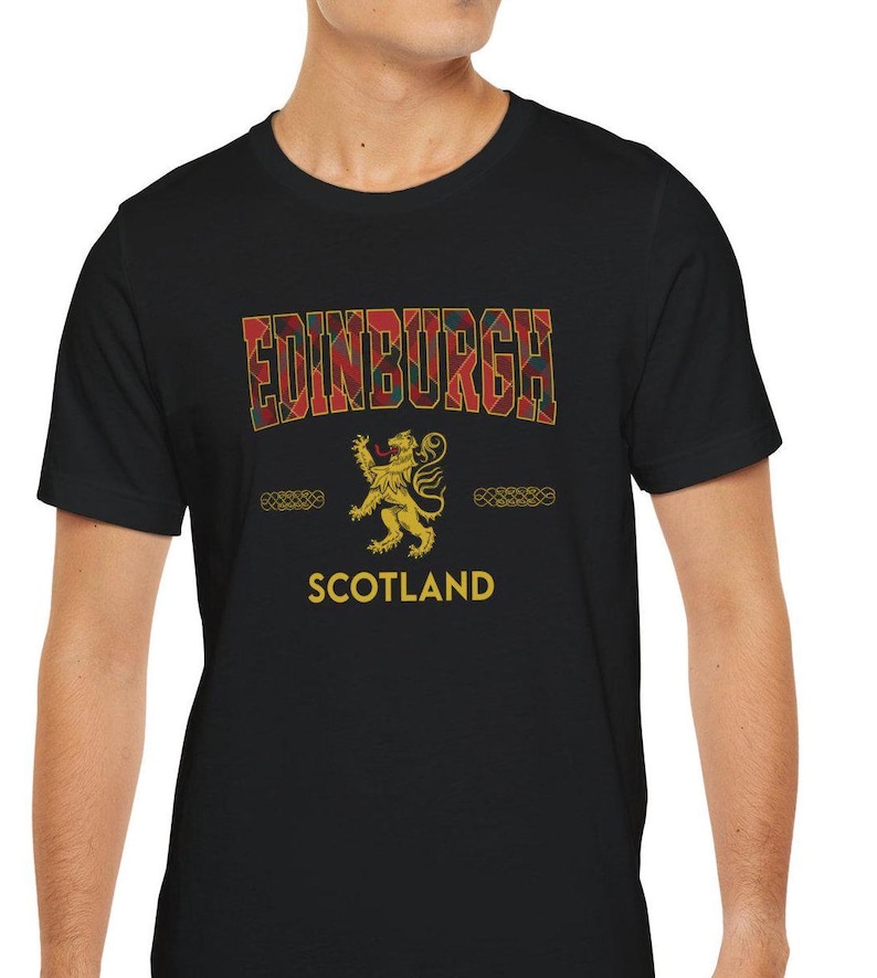 Puede incluir: Camiseta negra con la palabra "EDINBURGH" en un dise&ntilde;o de cuadros, un emblema de le&oacute;n dorado y la palabra "SCOTLAND" en dorado. La camiseta tiene cuello redondo y mangas cortas.
