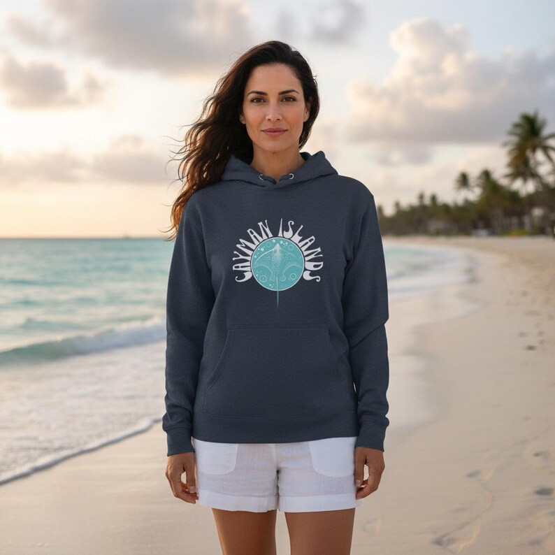 May include: A navy blue hoodie with the text "CAYMAN ISLANDS" in white, surrounding a light blue graphic of a stingray. The hoodie has a drawstring hood and a front pocket. The person is wearing white shorts on a beach.