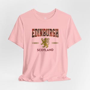 Puede incluir: Camiseta rosa claro con la palabra "EDINBURGH" en un dise&ntilde;o escoc&eacute;s. Debajo, un emblema de le&oacute;n dorado y la palabra "SCOTLAND". La camiseta tiene cuello redondo y mangas cortas.