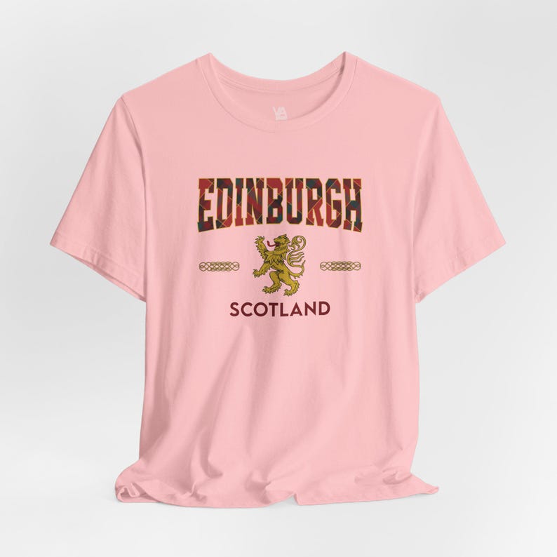 Puede incluir: Camiseta rosa claro con la palabra "EDINBURGH" en un dise&ntilde;o escoc&eacute;s. Debajo, un emblema de le&oacute;n dorado y la palabra "SCOTLAND". La camiseta tiene cuello redondo y mangas cortas.